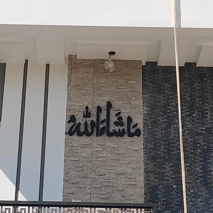Black Arabic calligraphy wall art mounted on a beige stone-textured panel, set against a modern white and dark gray building facade under a clear blue sky.